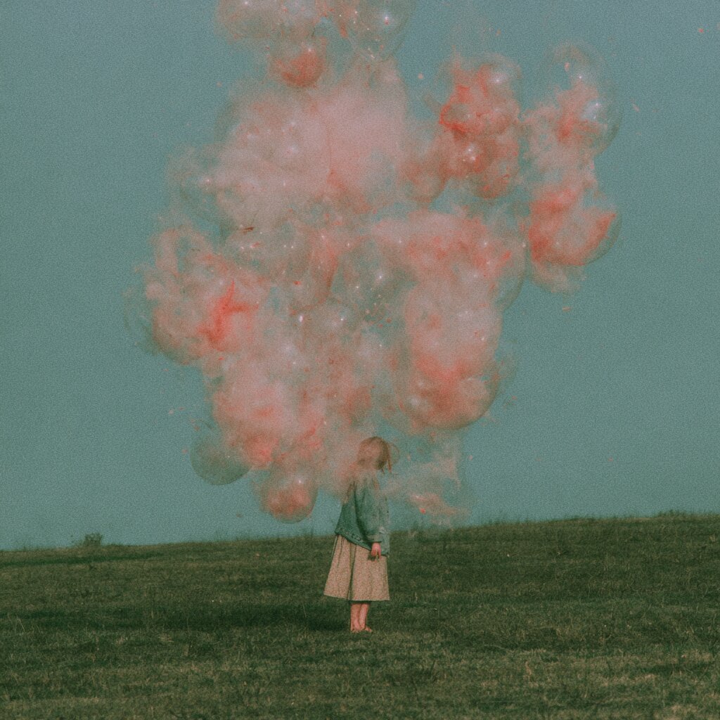 A nostalgic, filmic photograph of a young girl in a field looking up at a huge, suspended pink cloud that resembles a burst of bubbles